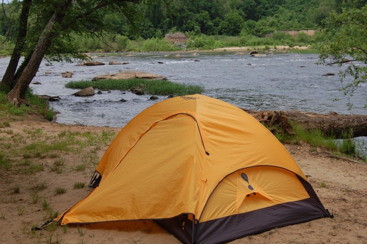 Yellow tent pitched by a riverside with trees and greenery in the background.