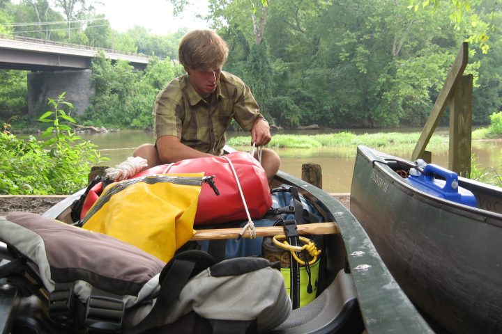 Person organizing gear in a canoe by a river, surrounded by trees, with a bridge in the background.